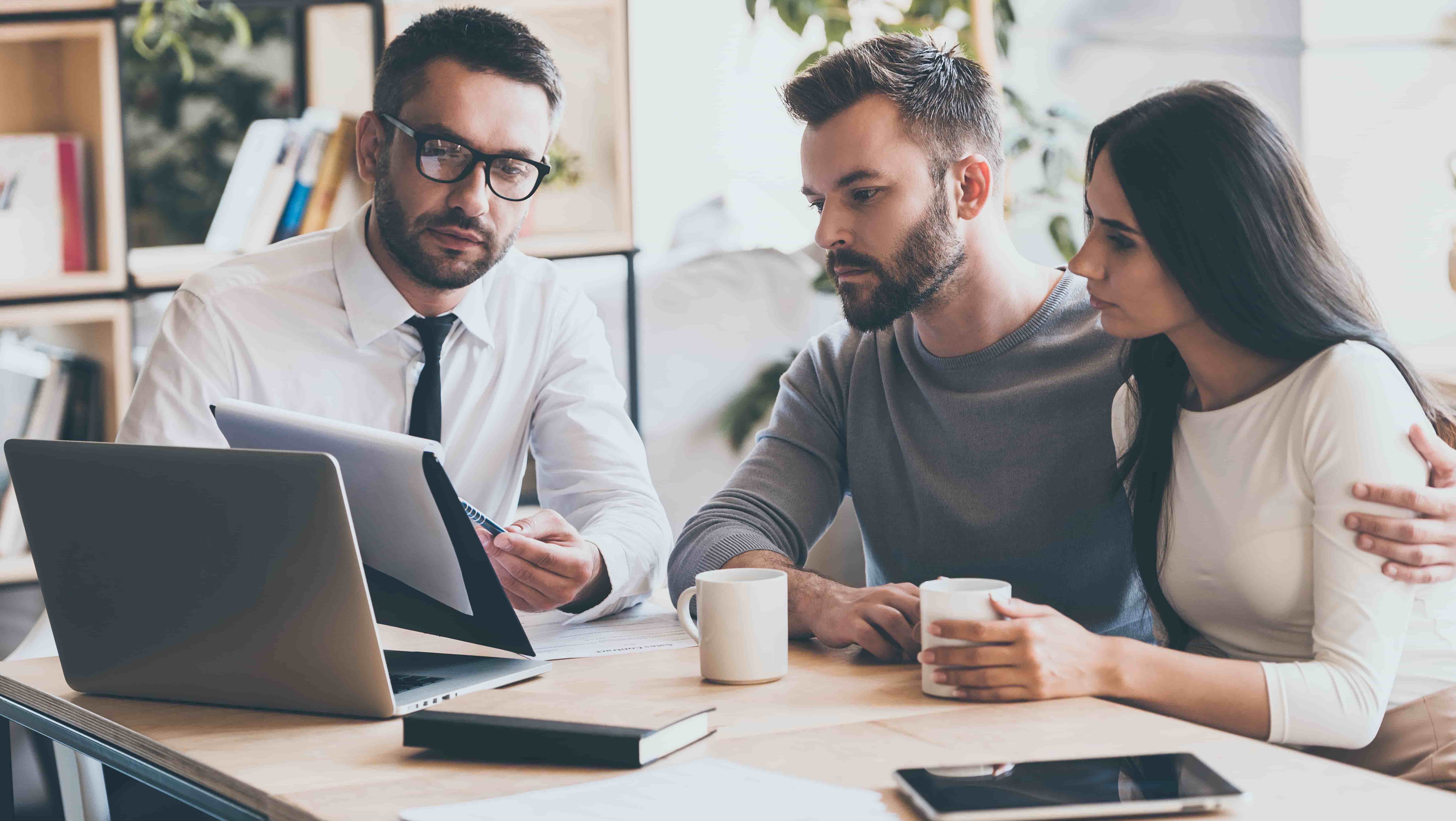 I need your signature here. Confident young man in shirt and tie holding some document and pointing it while sitting together with young couple at the desk in office